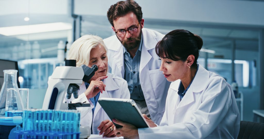 Three scientists in lab coats discuss information on a tablet, surrounded by lab equipment, including a microscope and test tubes, in a modern laboratory setting.