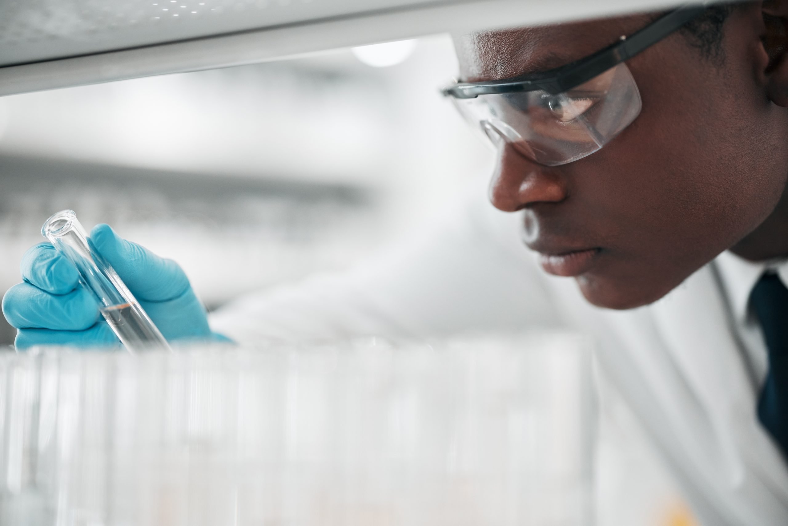 A scientist wearing safety goggles and blue gloves closely examines a test tube in a laboratory setting, surrounded by rows of empty test tubes.