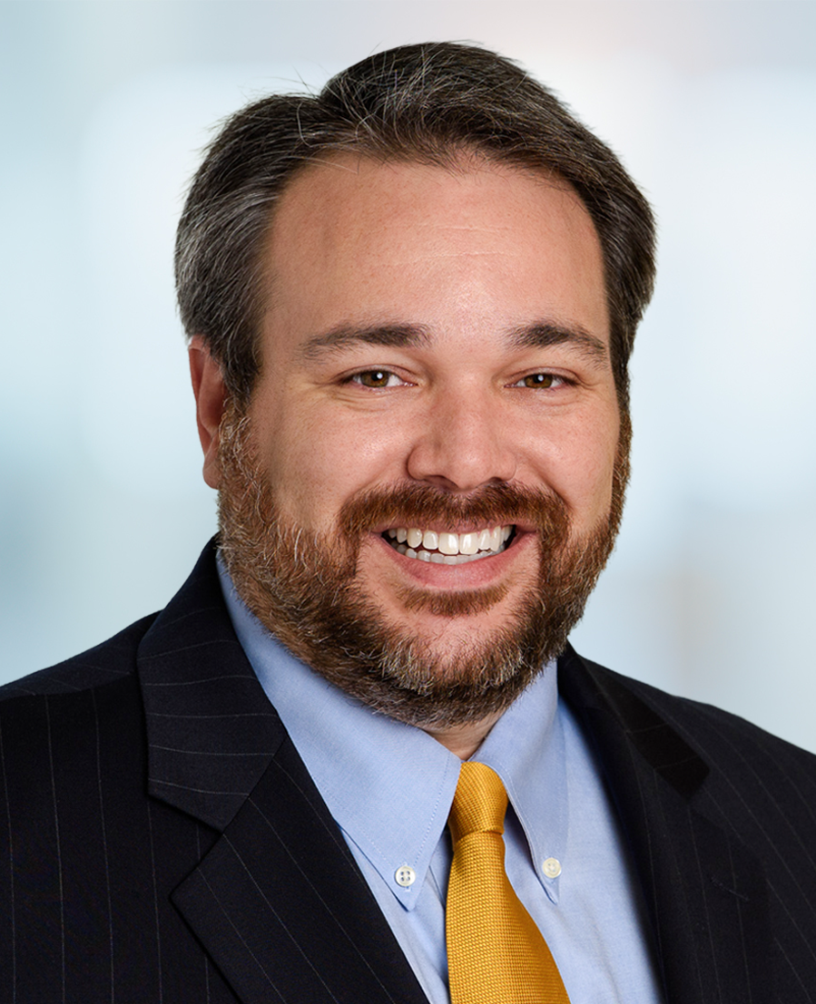 Smiling man with brown hair and beard, wearing a dark pinstripe suit, light blue shirt, and yellow tie, posed against a blurred light background.