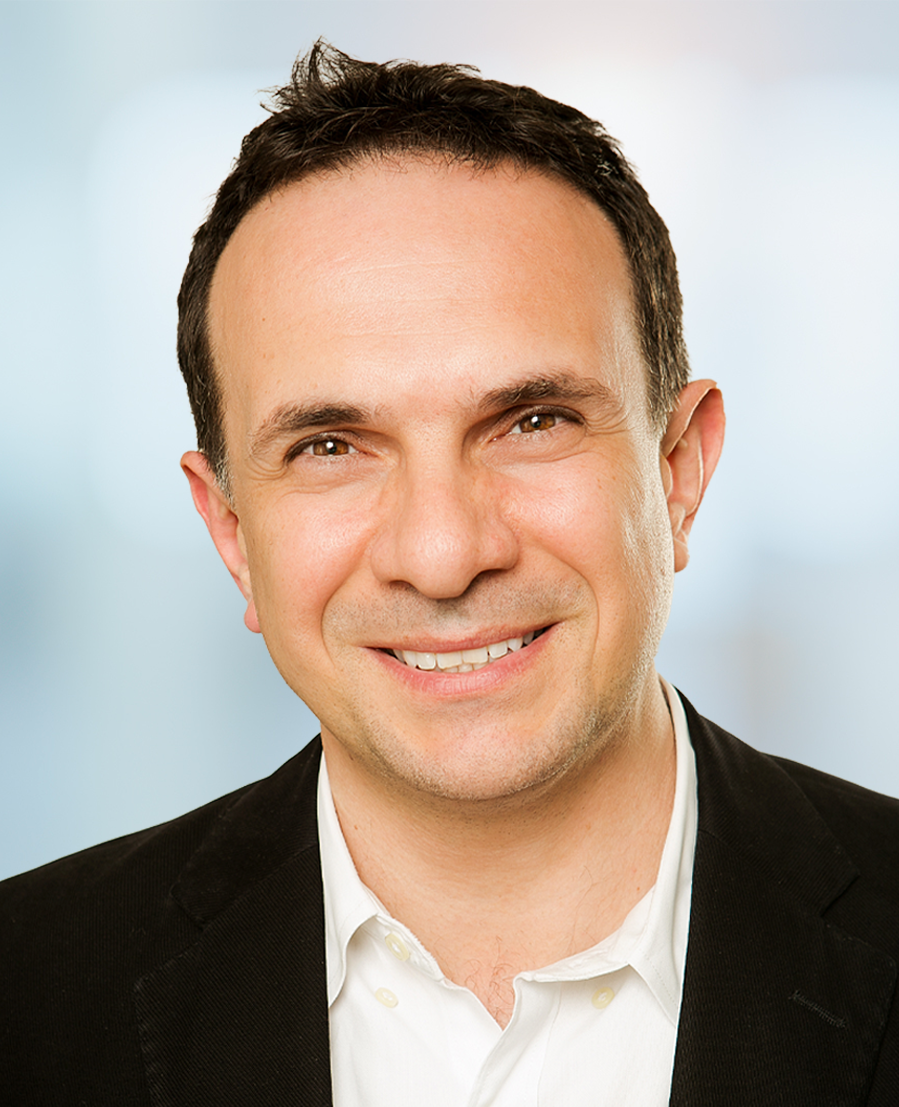 A man with short dark hair smiles at the camera. He is wearing a white shirt and a dark blazer. The background is softly blurred in shades of blue and white.