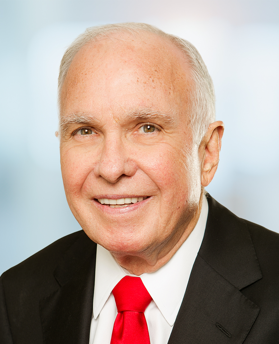 Smiling older man with short white hair, wearing a black suit, white shirt, and bright red tie, posing in front of a softly blurred blue and white background.