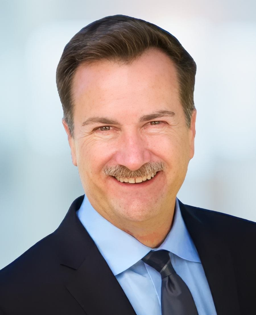 A man in a suit and tie smiles at the camera against a blurred light background.