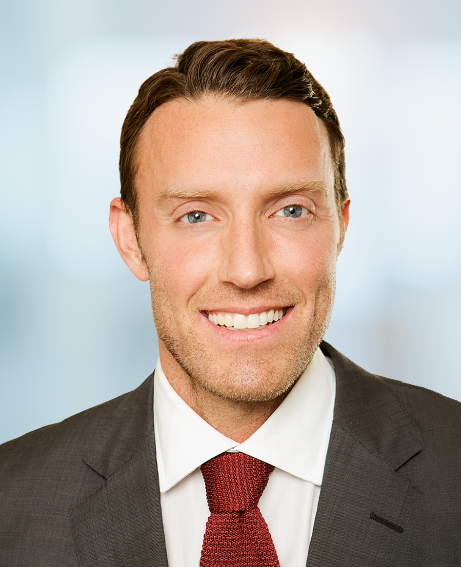 Smiling man wearing a gray suit, white shirt, and red tie, posing against a blurred light blue background.