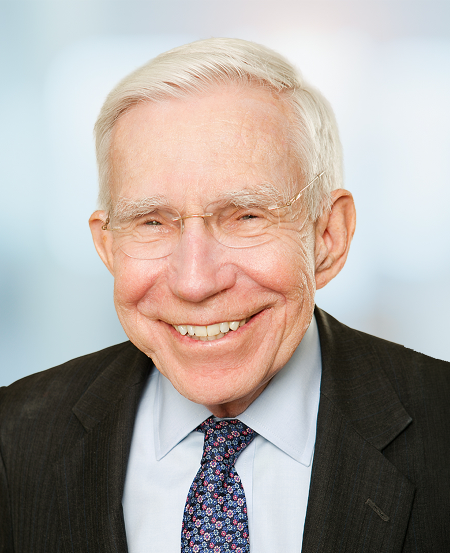 Smiling older man with short white hair, wearing glasses, a dark suit, a light blue shirt, and a floral-patterned tie, against a blurred light background.