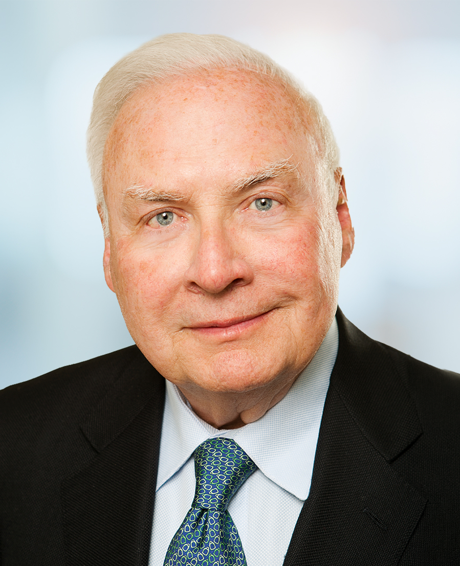 An older man with white hair, wearing a dark suit, white shirt, and patterned tie, poses for a formal portrait against a softly blurred background.
