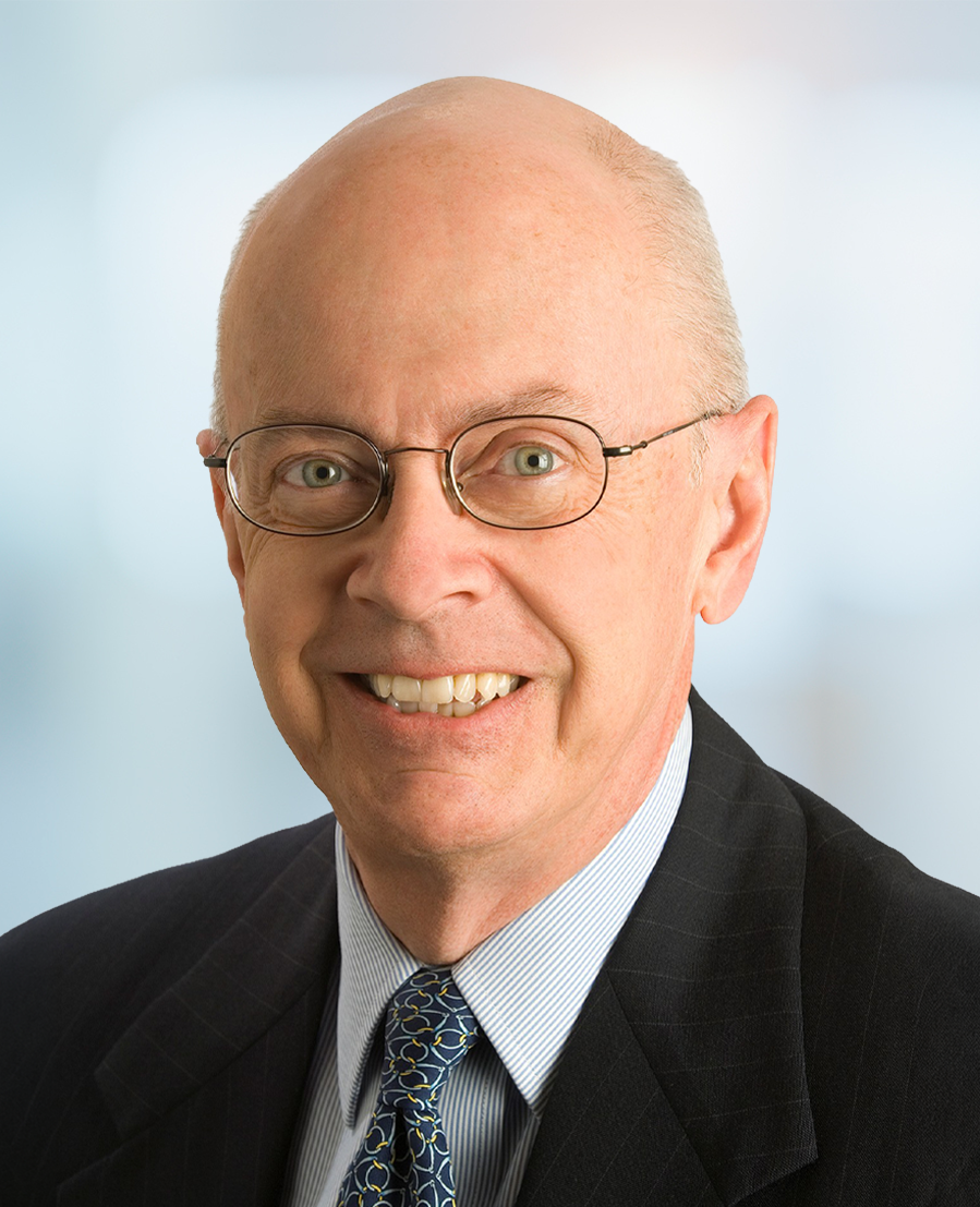 A smiling older man with glasses and a bald head, wearing a dark suit, striped shirt, and a patterned tie, poses in front of a softly blurred background.