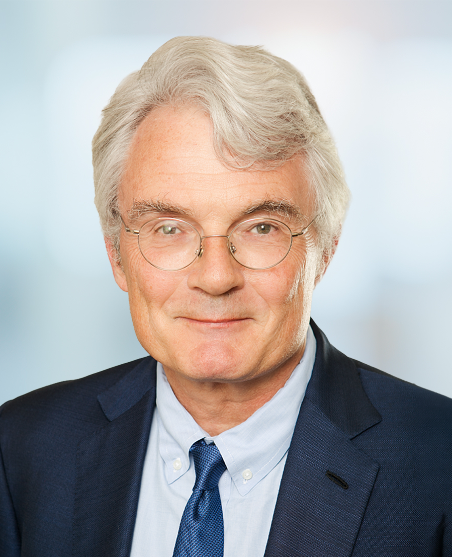 An older man with gray hair, glasses, and a blue suit with a tie, smiling slightly against a softly blurred light background.