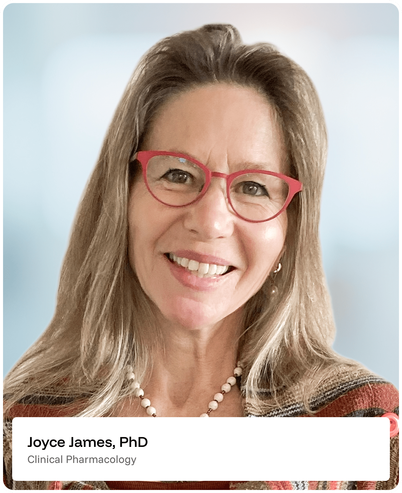 Smiling woman with long light brown hair and red glasses, wearing a plaid top and beaded necklace. Text reads: Joyce James, PhD. Clinical Pharmacology. Background is softly blurred.