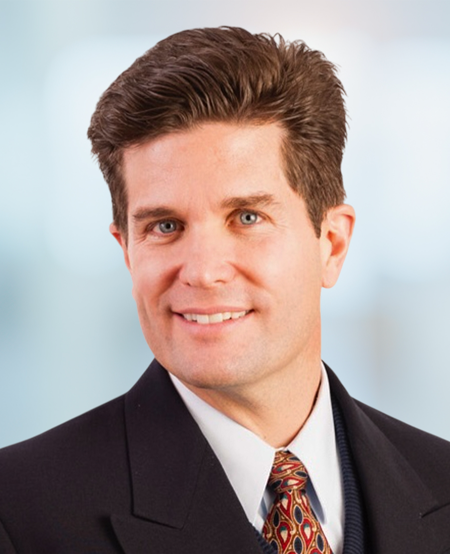 A man with short brown hair wearing a dark suit, white shirt, and patterned tie smiles at the camera. The background is blurred in light blue tones.