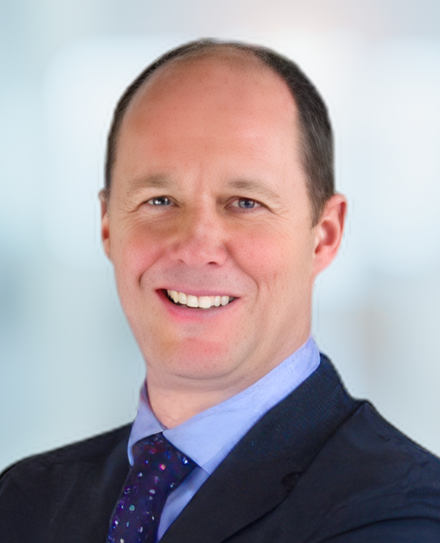 Smiling man in a dark suit and purple tie with a light blue shirt, standing against a softly blurred light background.