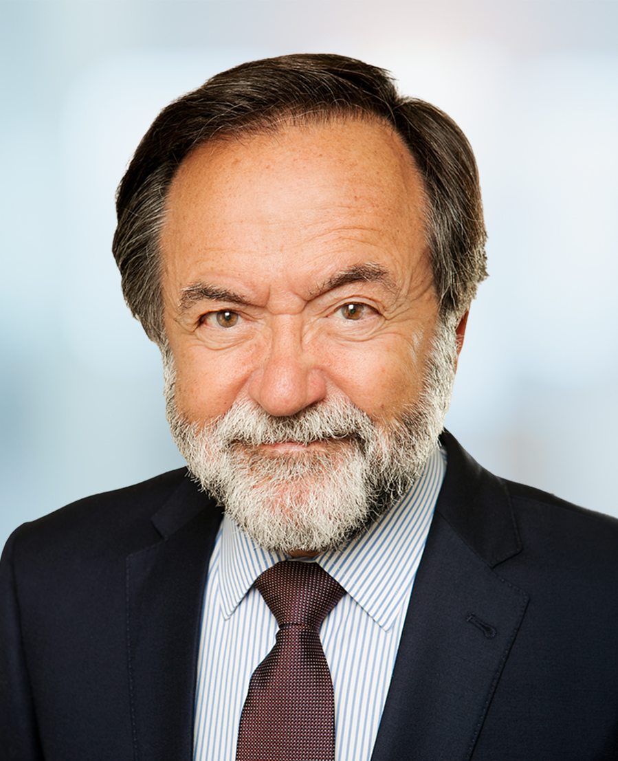 An older man with gray hair and a beard, wearing a dark suit, striped shirt, and maroon tie, smiles at the camera against a blurred light background.