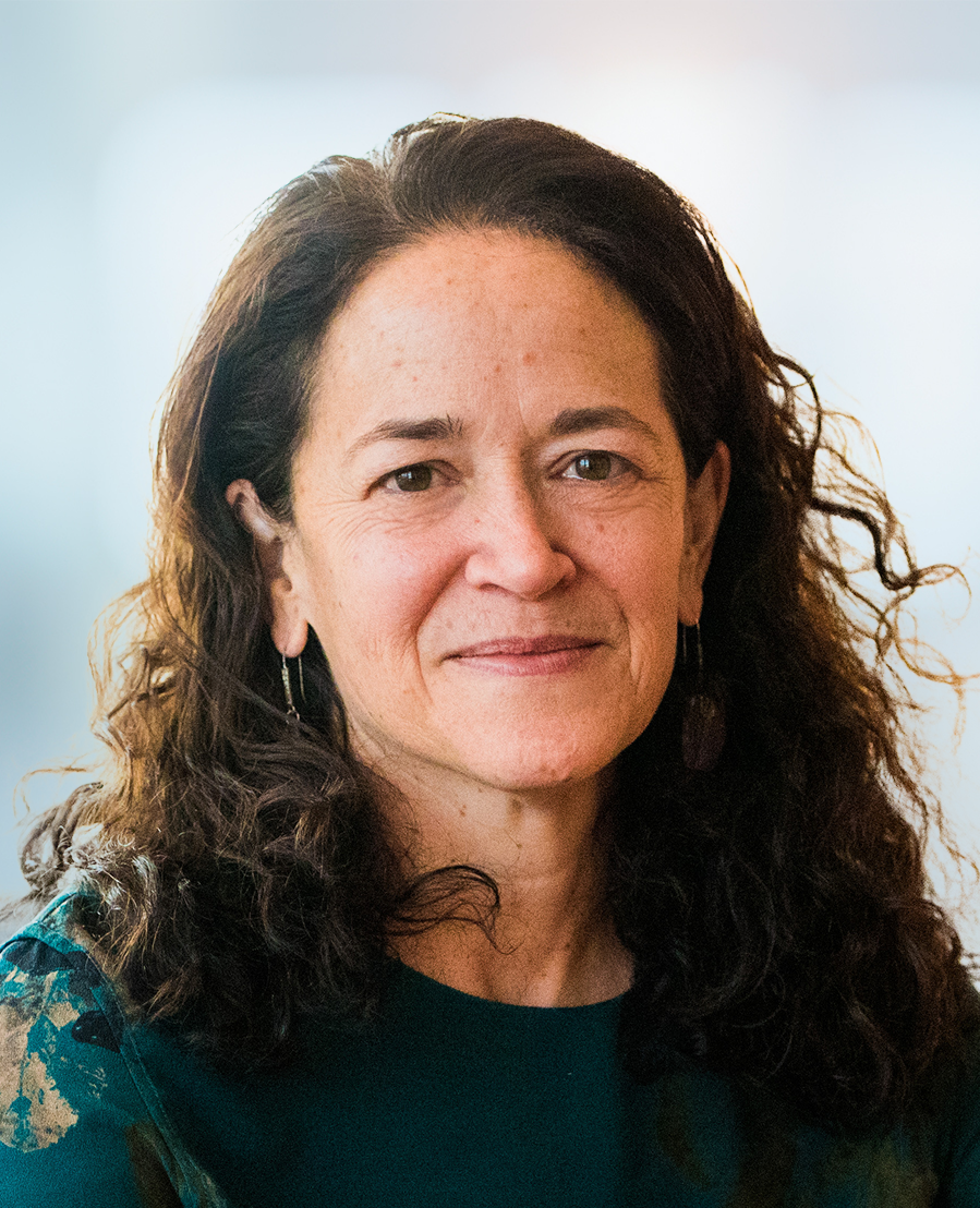 A woman with long, curly brown hair and light skin smiles gently at the camera, wearing a green top and hoop earrings. The background is softly blurred.