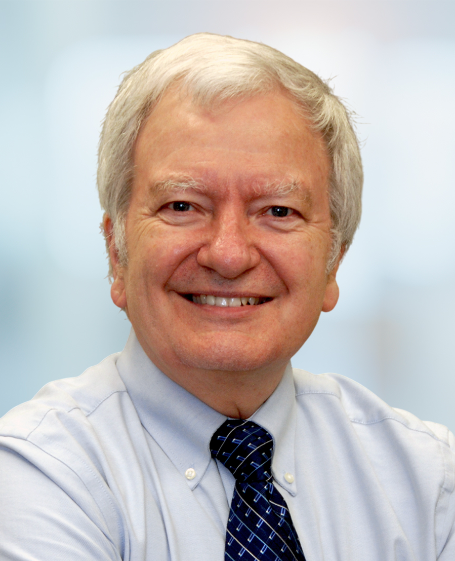 Smiling older man with short white hair, wearing a light blue collared shirt and a dark blue patterned tie, photographed against a blurred office background.