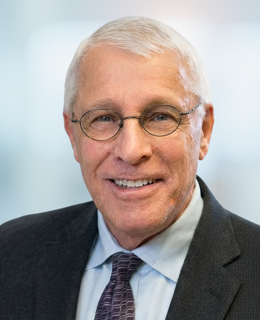 An older man with short gray hair, glasses, and a friendly smile, wearing a dark suit, light blue shirt, and patterned tie, posed against a blurred, light-colored background.