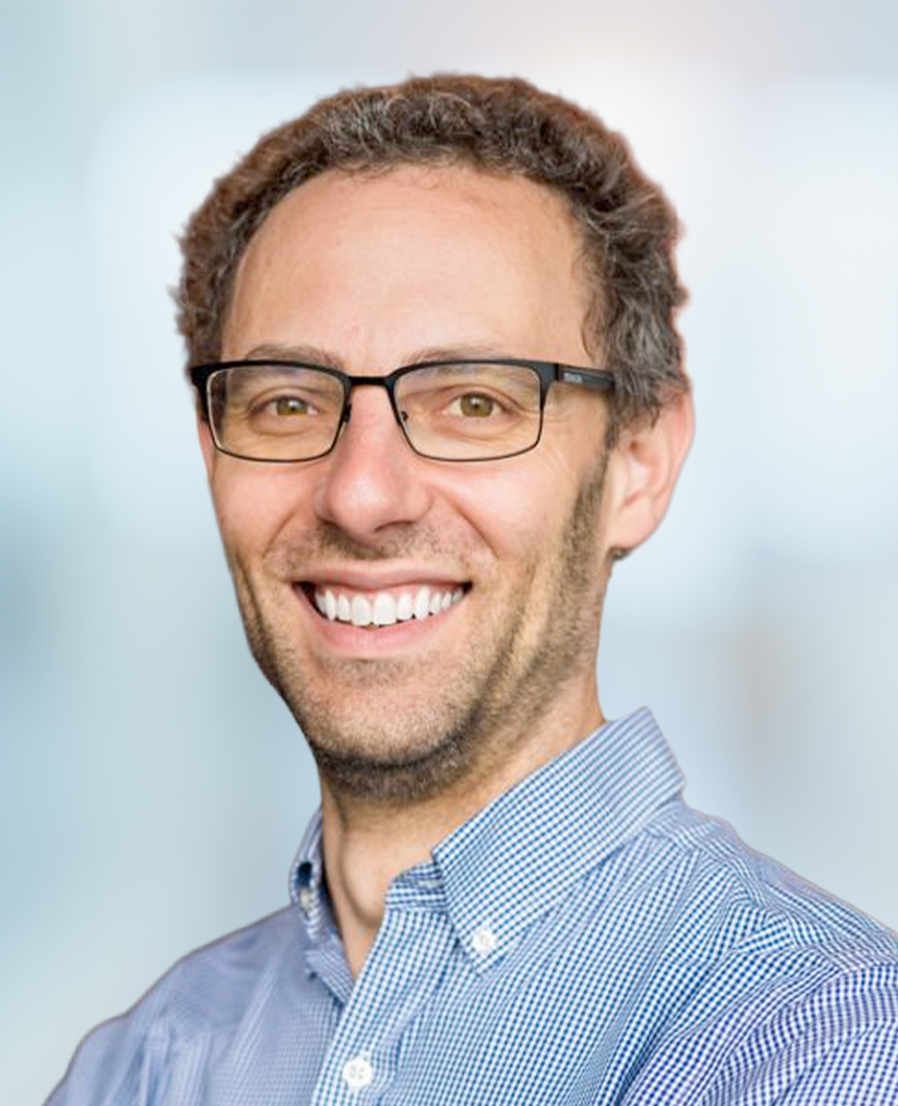 Smiling man with curly hair, glasses, and a checked button-up shirt, standing in front of a blurred light blue background.