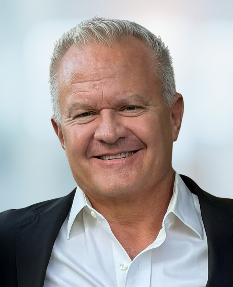 Smiling man with short gray hair wearing a white collared shirt and a black jacket, standing against a blurred light background.