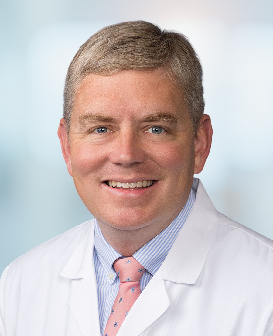 Smiling man with short blond hair wearing a white medical coat, blue striped shirt, and pink tie with small blue prints, posed in front of a blurred light background.